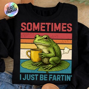 Op de afbeelding: Zwarte sweatshirt met een afbeelding van een groene kikker die een gele mok vasthoudt, zittend op een waterlelieblad. De tekst luidt "SOMETIMES I JUST BE FARTIN'" boven en onder de kikker, met een retro zonsondergang achtergrond.