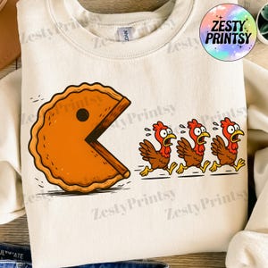 May include: Cream-colored sweatshirt featuring a humorous graphic of a pie chasing three running chickens. The pie has a Pac-Man-like design. The text "Zesty Printsy" is in a circle on the upper right.