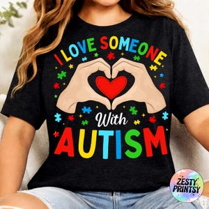 May include: Black t-shirt with the text "I LOVE SOMEONE WITH AUTISM" in colorful letters. The design features hands forming a heart around a red heart, with puzzle pieces scattered around.