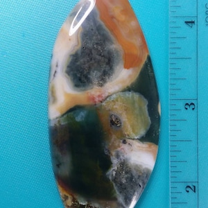 May include: A polished, oval-shaped, green, brown, and white agate stone with a ruler for scale. The stone is approximately 3 inches long.