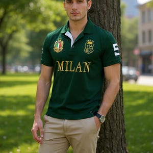 May include: A dark green short-sleeved polo shirt featuring the word MILAN in gold lettering across the chest, a crest with the Italian flag on the right side, a shield emblem on the left, and the number 5 printed in white on the left sleeve.