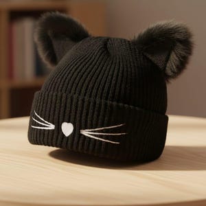 May include: Black knitted beanie hat with embroidered white cat whiskers and a heart-shaped nose. The hat has faux fur ears. A fun winter accessory for cat lovers.
