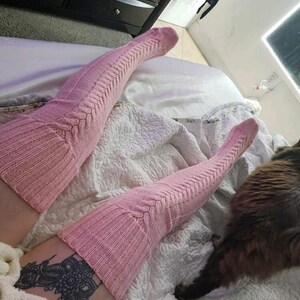 May include: A pair of light pink, cable-knit thigh-high socks. The socks have a ribbed texture at the top and bottom, with a cable-knit design running down the length. The socks are worn on a bed with a white comforter.