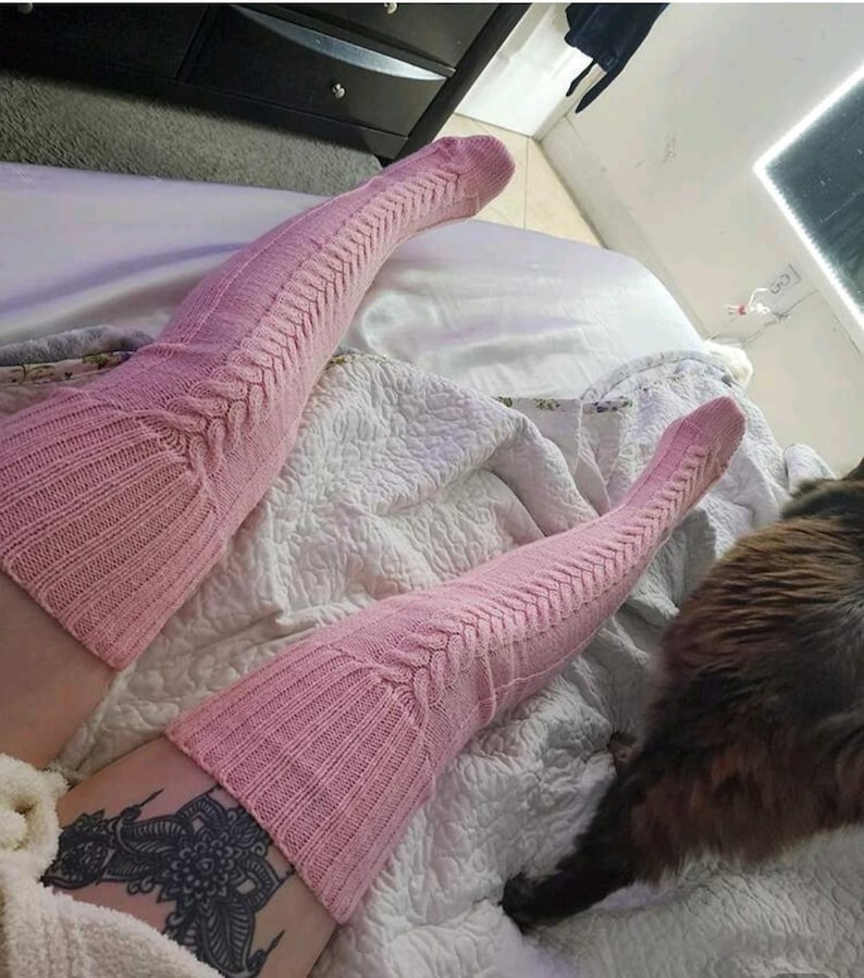 May include: A pair of light pink, cable-knit thigh-high socks. The socks have a ribbed texture at the top and bottom, with a cable-knit design running down the length. The socks are worn on a bed with a white comforter.
