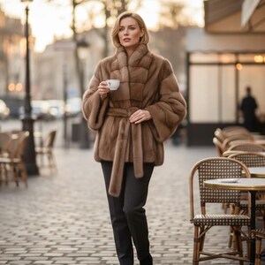 Peut inclure: Une veste en fourrure marron clair avec un col montant, des manches larges et une ceinture à nouer assortie à la taille, portée avec un pantalon sombre. La veste présente un design à panneaux horizontaux et est portée sur un pantalon simple.