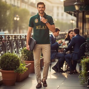 May include: A dark green short-sleeved polo shirt with the word MILAN printed in gold letters across the chest, paired with beige trousers and brown loafers. The shirt features a small crest emblem on the chest and a white inner collar detail.