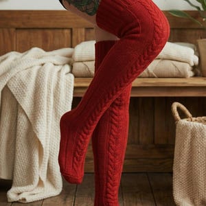 May include: Red cable-knit over-the-knee socks. The socks are a vibrant red color and feature a ribbed texture on the leg and a cable-knit design on the sides. The socks are made of a thick, warm material, perfect for cold weather.