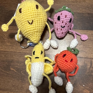 May include: Four handmade crochet fruit keychains: a yellow lemon, a pink strawberry, a yellow banana, and an orange tomato. Each fruit has a smiling face, arms, legs, and white feet. The lemon has a silver keychain.