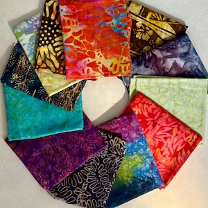 May include: A collection of folded fabric squares in various colors and patterns. The fabrics include vibrant hues of red, blue, purple, and green, with designs such as floral, batik, and abstract motifs. The arrangement is circular, showcasing the diverse textiles.