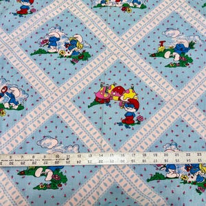 May include: Light blue fabric with a repeating pattern of Smurfs, mushrooms, and flowers. The design is set within a white lattice pattern. The fabric is measured with a ruler.