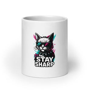 Könnte beinhalten: Weißer Keramikbecher mit einer stilisierten schwarz-weißen Katze, die eine Sonnenbrille und ein Nietenhalsband trägt. Das Design zeigt den Text „STAY SHARP“ in fetter Schrift, mit rosa und türkisfarbenen Akzenten.