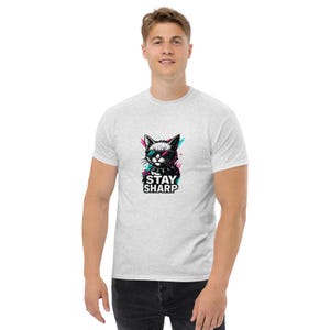Könnte beinhalten: Hellgraues T-Shirt mit einer Grafik einer Katze mit Sonnenbrille und dem Text "STAY SHARP". Die Katzengrafik hat rosa, blaue und schwarze Akzente. Das Shirt ist ein klassischer Rundhalsausschnitt.