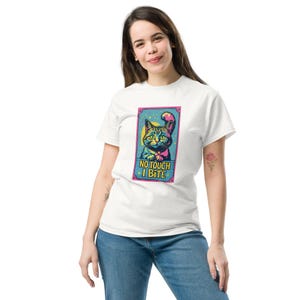 Könnte beinhalten: Weißes T-Shirt mit einer bunten Grafik einer Katze und dem Text "NO TOUCH I BITE". Die Katzenillustration hat einen gelben Hintergrund und einen rosa und blauen Rahmen. Die Katze hat blaues und grünes Fell.