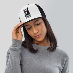 May include: A black and white trucker hat with a white front panel featuring a graphic of a cat with pink and blue accents and the text "STAY SHARP". The hat has a black brim and mesh back.