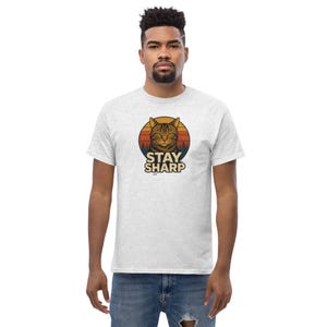 Könnte beinhalten: Hellgraues T-Shirt mit einem kreisförmigen Grafikdesign eines Katzenkopfes in einem Sonnenuntergang. Der Text "STAY SHARP" ist darunter gedruckt. Das Shirt ist aus weichem Material.