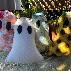 May include: Four handmade ghost-shaped plush dolls. Each ghost has black oval eyes and is made from different patterned fabrics, including floral and sunflower prints. Some are adorned with bows and flowers.