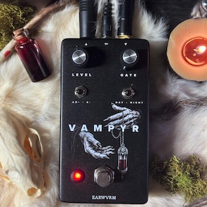 May include: A black guitar effects pedal labeled VAMPYR and EARWVRM, featuring two silver knobs for level and gate, two toggle switches, and a graphic of pale hands holding a vial pendant. A red LED indicator light is illuminated near the metal footswitch.