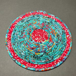 May include: A round, spiral-patterned trivet or coaster. The center is red, surrounded by a turquoise and patterned fabric, and edged with a red border. The item is made of tightly woven fabric.