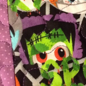 Puede incluir: Primer plano de una tela con tem&aacute;tica de Halloween con un dise&ntilde;o de patchwork. La tela presenta un monstruo de Frankenstein con piel verde, pelo morado y ojos rojos. La tela tambi&eacute;n tiene un estampado de lunares morados.