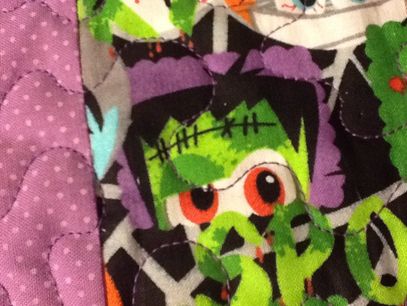 Puede incluir: Primer plano de una tela con tem&aacute;tica de Halloween con un dise&ntilde;o de patchwork. La tela presenta un monstruo de Frankenstein con piel verde, pelo morado y ojos rojos. La tela tambi&eacute;n tiene un estampado de lunares morados.