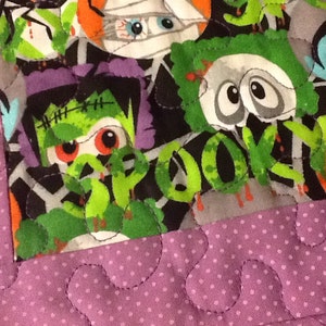 Puede incluir: Primer plano de un edred&oacute;n con tem&aacute;tica de Halloween con varios monstruos de dibujos animados como Frankenstein, momias y vampiros. El edred&oacute;n tiene un borde morado con lunares y un patr&oacute;n de puntadas onduladas. La tela de los monstruos es gris, verde, naranja y morada.