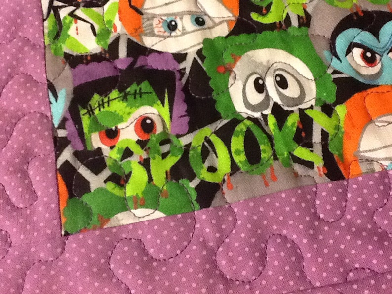 Puede incluir: Primer plano de un edred&oacute;n con tem&aacute;tica de Halloween con varios monstruos de dibujos animados como Frankenstein, momias y vampiros. El edred&oacute;n tiene un borde morado con lunares y un patr&oacute;n de puntadas onduladas. La tela de los monstruos es gris, verde, naranja y morada.