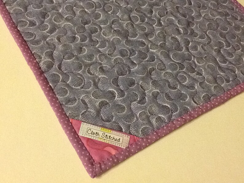 Puede incluir: Textil acolchado con un patr&oacute;n gris y blanco de c&iacute;rculos entrelazados. El borde est&aacute; adornado con una tela rosa de lunares y un borde rosa. La etiqueta dice "Cloth Stitched".