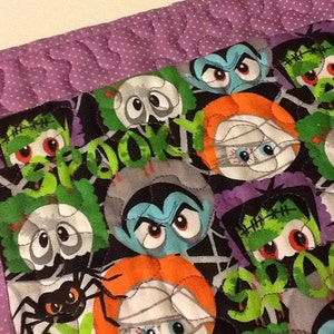 Puede incluir: Un edred&oacute;n con tem&aacute;tica de Halloween con monstruos de dibujos animados, ara&ntilde;as y la palabra "BOO!" en verde. El edred&oacute;n tiene un fondo negro con un borde morado y un patr&oacute;n de puntos. Los monstruos incluyen Frankenstein, vampiros y momias.