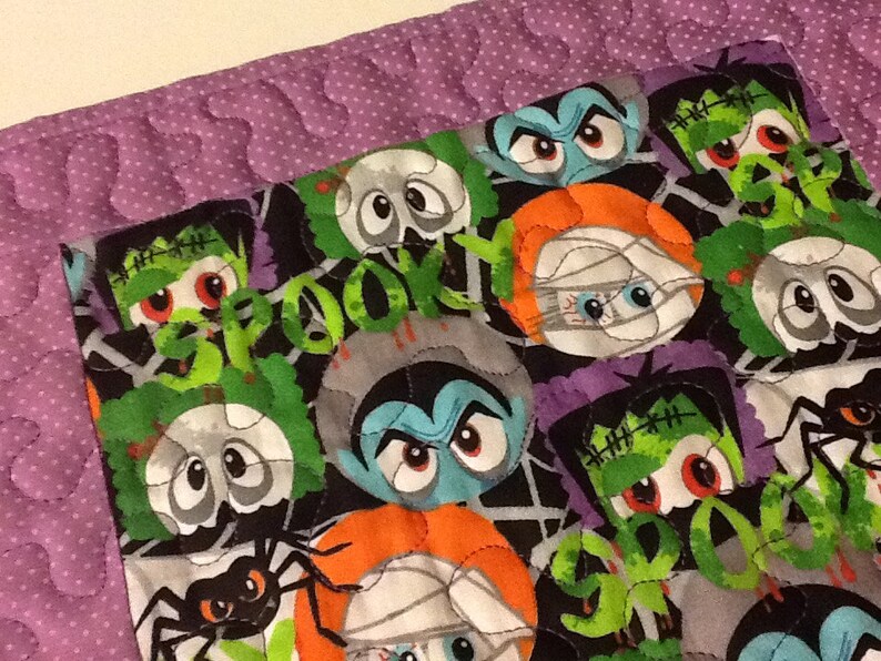 Puede incluir: Un edred&oacute;n con tem&aacute;tica de Halloween con monstruos de dibujos animados, ara&ntilde;as y la palabra "BOO!" en verde. El edred&oacute;n tiene un fondo negro con un borde morado y un patr&oacute;n de puntos. Los monstruos incluyen Frankenstein, vampiros y momias.