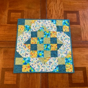 May include: A square, handmade quilt with a patchwork design. The quilt features a teal border and a central pattern of squares and floral shapes in shades of blue, yellow, and green. The quilt is approximately 12 inches square.