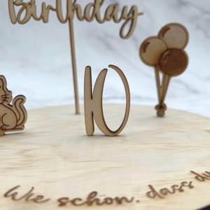 Op de afbeelding: Een ronde houten taarttopper met het woord Birthday in script, het getal 10, een katfiguur en drie ballonnen. De basis is gegraveerd met de Duitse tekst Wie schon, dass du geboren bist in een cursief lettertype, alles uitgevoerd in licht hout.