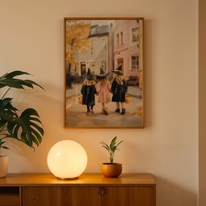 May include: A framed painting depicts three children in witch costumes walking down a street. The children are holding hands and carrying jack-o'-lantern buckets. The painting is displayed above a wooden cabinet with a round lamp and a potted plant.