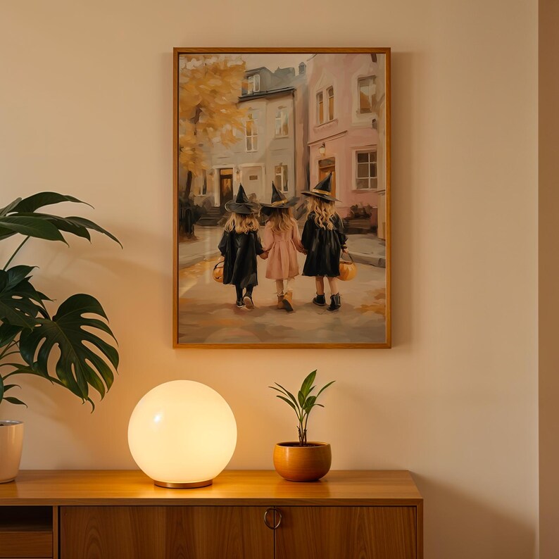 May include: A framed painting depicts three children in witch costumes walking down a street. The children are holding hands and carrying jack-o'-lantern buckets. The painting is displayed above a wooden cabinet with a round lamp and a potted plant.