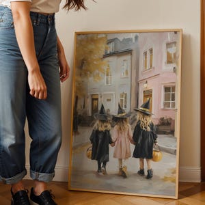 May include: A framed print depicts three young children dressed as witches, walking down a street. The children wear black cloaks and hats, with one in a pink dress. They carry pumpkin-shaped baskets, set against a backdrop of European-style buildings.