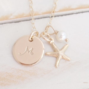 May include: Gold necklace with a round charm engraved with the letter "M", a small starfish charm, and a pearl charm.