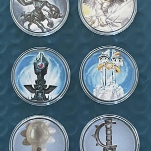 May include: A collection of six circular game pieces featuring various fantasy characters and objects. The pieces have detailed illustrations in a variety of colors, including silver, blue, and gold. The designs include a centaur-like creature, an angel, and a weapon.