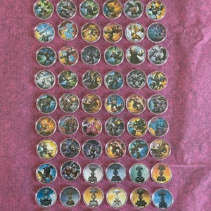 May include: A collection of circular game tokens with various character illustrations. The tokens have a metallic silver border and are arranged on a pink surface. The illustrations feature a variety of colorful fantasy characters.