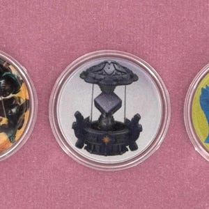 May include: Three round, clear plastic tokens with colorful illustrations. One depicts a warrior with a bow, another a stylized fountain, and the last a blue treasure chest on a yellow background. The tokens are set against a pink background.