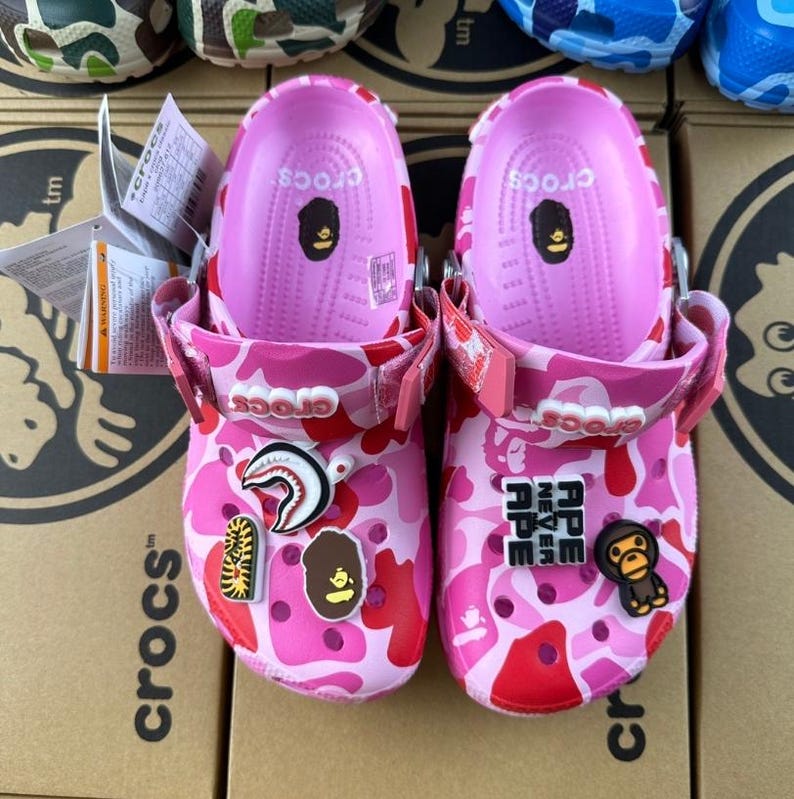 May include: Pink Crocs clogs with a camouflage design in pink and red hues. The clogs are adorned with various charms, such as a shark, tiger, and a cartoon ape. The interior is a solid pink, and the clogs have a strap for a secure fit.