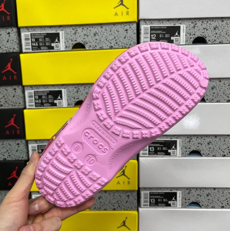May include: A pink Crocs shoe sole, size 8/10, with a textured pattern. The shoe is made in China. The background shows stacked shoe boxes with the word "AIR" and the numbers "12" and "13".