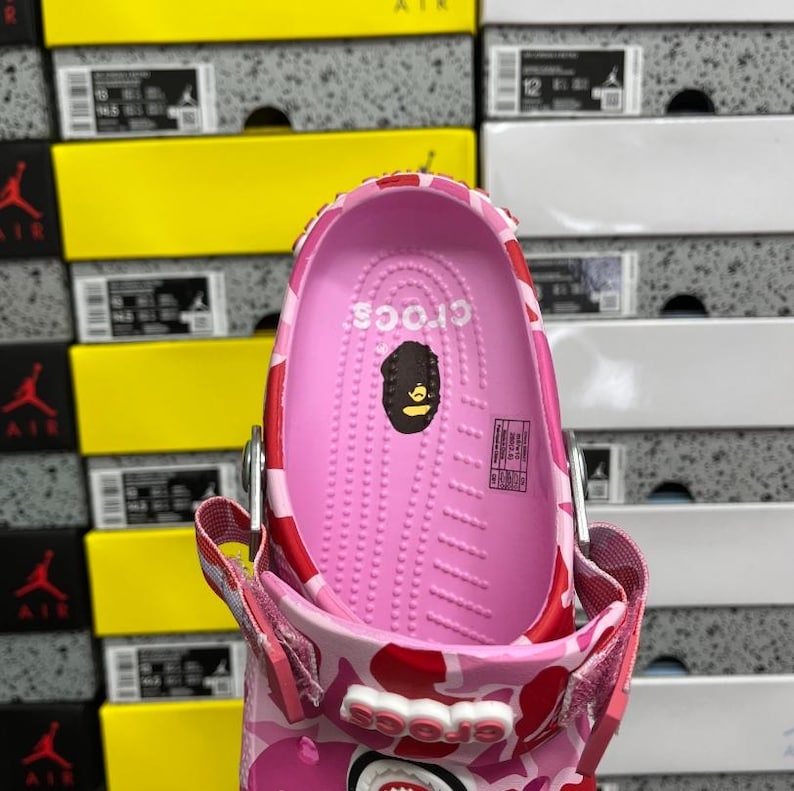 May include: Pink Crocs shoe with a camouflage pattern in red and white. The interior is pink with the Crocs logo and a graphic. The shoe is set against a backdrop of shoe boxes in yellow and white.