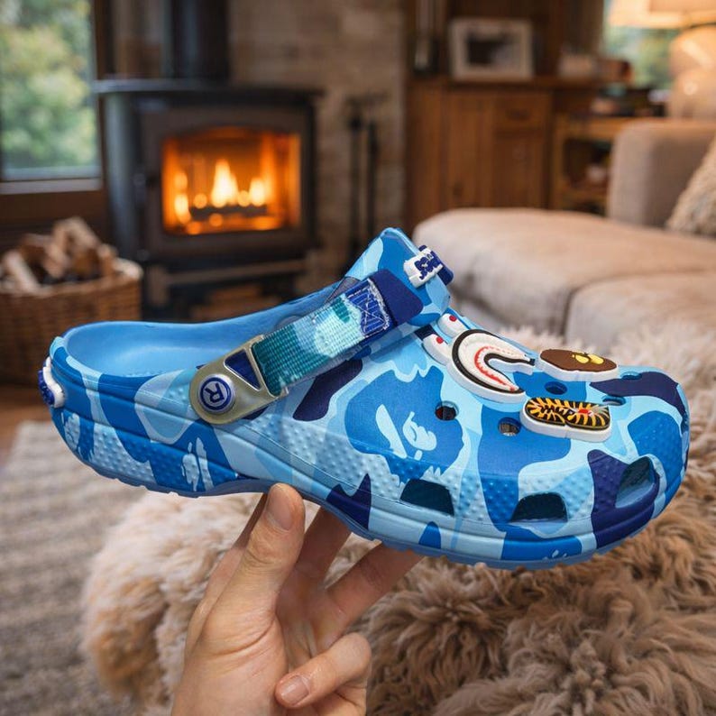 May include: Blue camouflage-patterned clog with decorative charms. The shoe features a pivoting heel strap and a blue and white camouflage design. The clog is held by a hand, showcasing the shoe's details and design.