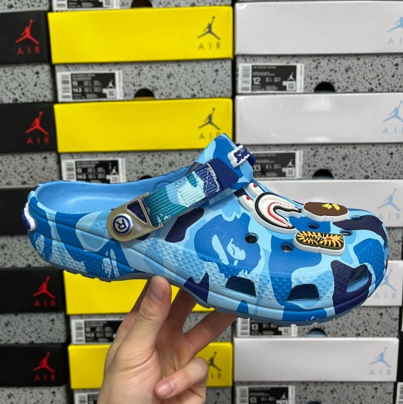 May include: A blue camouflage clog with a pivoting heel strap. The shoe has decorative charms, including a shark, sushi, and a tiger. The shoe is held by a hand, with shoe boxes in the background.