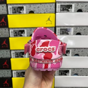 May include: Pink Crocs clogs with a red and pink camouflage design. The word "crocs" is printed in white. The sole features the text "A BATHING APE" in white. The clogs are held in front of a background of shoe boxes.