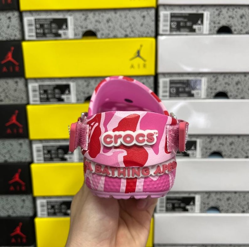 May include: Pink Crocs clogs with a red and pink camouflage design. The word "crocs" is printed in white. The sole features the text "A BATHING APE" in white. The clogs are held in front of a background of shoe boxes.