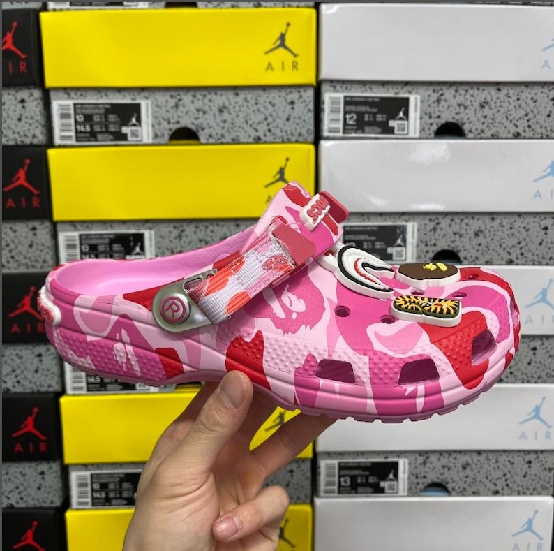 May include: Pink camouflage clogs with a pivoting heel strap and decorative charms. The clogs feature a pink and red camouflage pattern and are held by a hand. The background shows stacked shoe boxes.