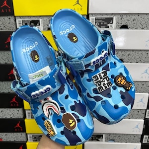 May include: A pair of blue camouflage Crocs sandals with various decorative charms. The sandals feature a blue and white pattern, with the word "Crocs" in white on the straps. Additional charms include a shark, a monkey, and the text "APE".