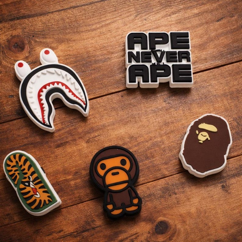 May include: A selection of shoe charms with diverse designs. Includes a white shark head with red and black details, a tiger on a green background, a brown ape, a brown ape head, and a black and white text that says "APE NEVER APE".