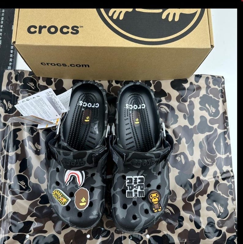 May include: Two black Crocs clogs with a grey camouflage design. The clogs have decorative patches and are presented with their original box. The box displays the Crocs logo and website address.
