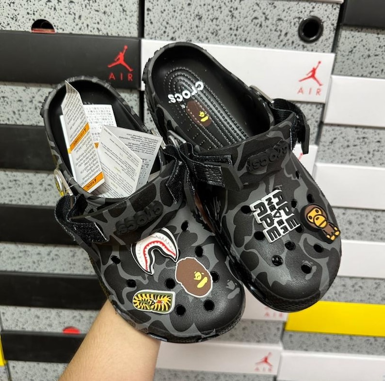 May include: A pair of black and grey camouflage Crocs clogs with decorative charms. The clogs have adjustable straps and the word "Crocs" is visible on the inner sole. Several decorative charms are attached to the clogs.